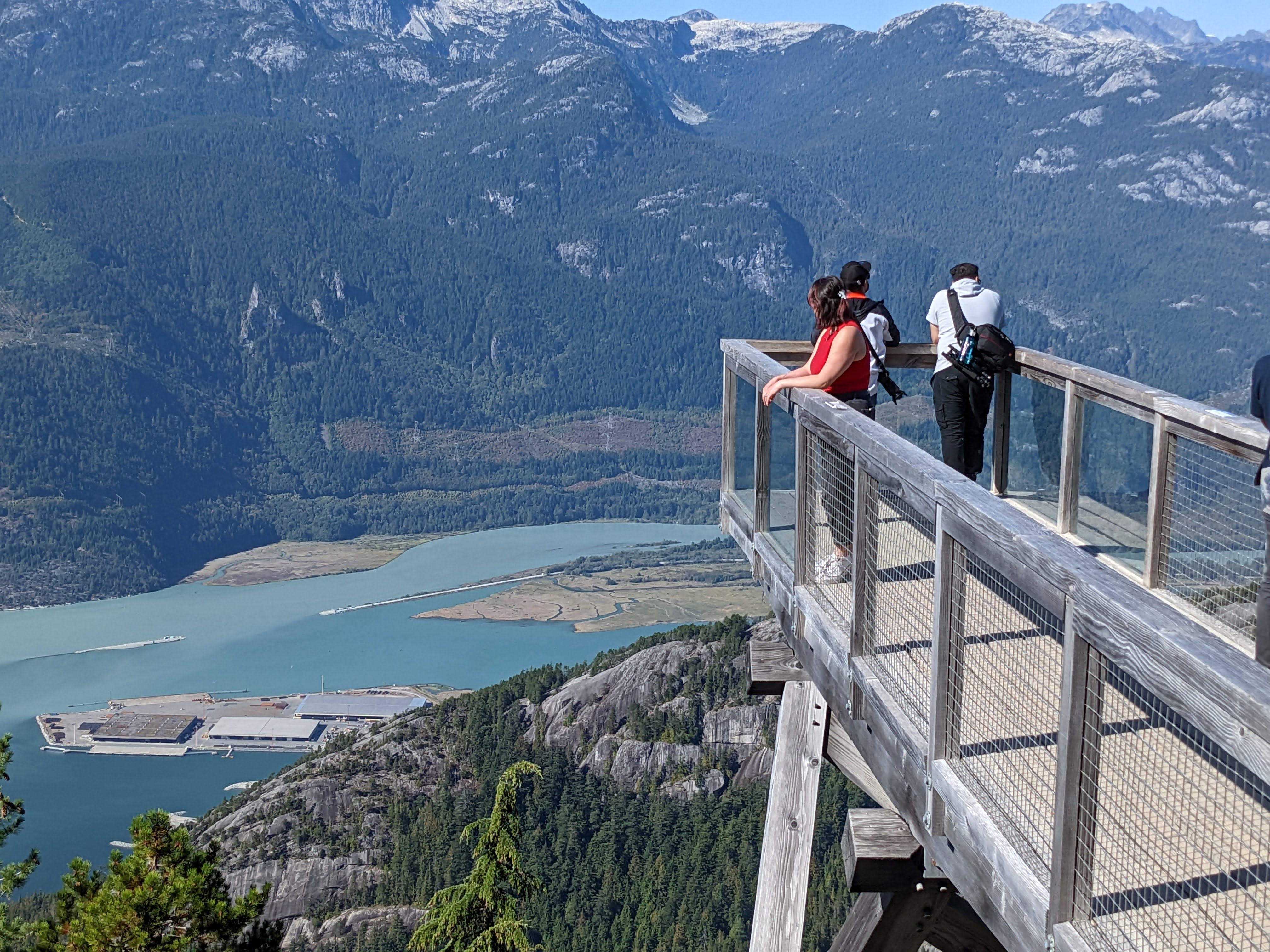 some people standing on a wooden overlook. They're looking out into a mountainous valley with a river cutting through the middle.  